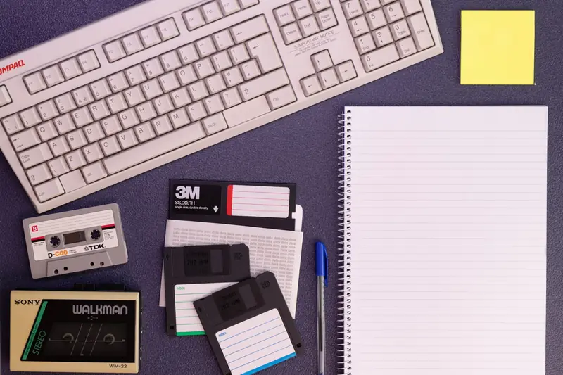 Vintage office setup featuring a Compaq keyboard, floppy disks, cassette tape, and notepad