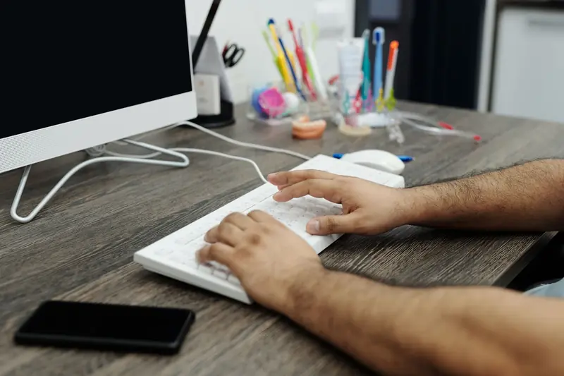 Close-up of hands typing on a white keyboard at a wooden desk in an office setting with accessories